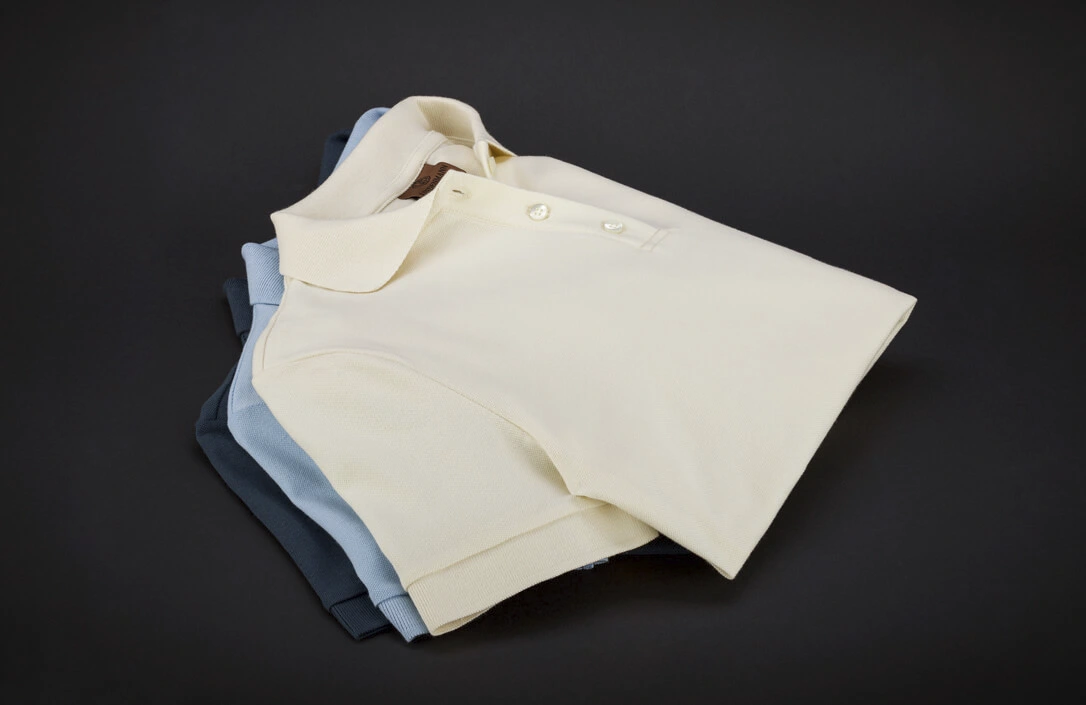 Drei gefaltete Poloshirts in Creme, Blau und Schwarz auf dunklem Hintergrund