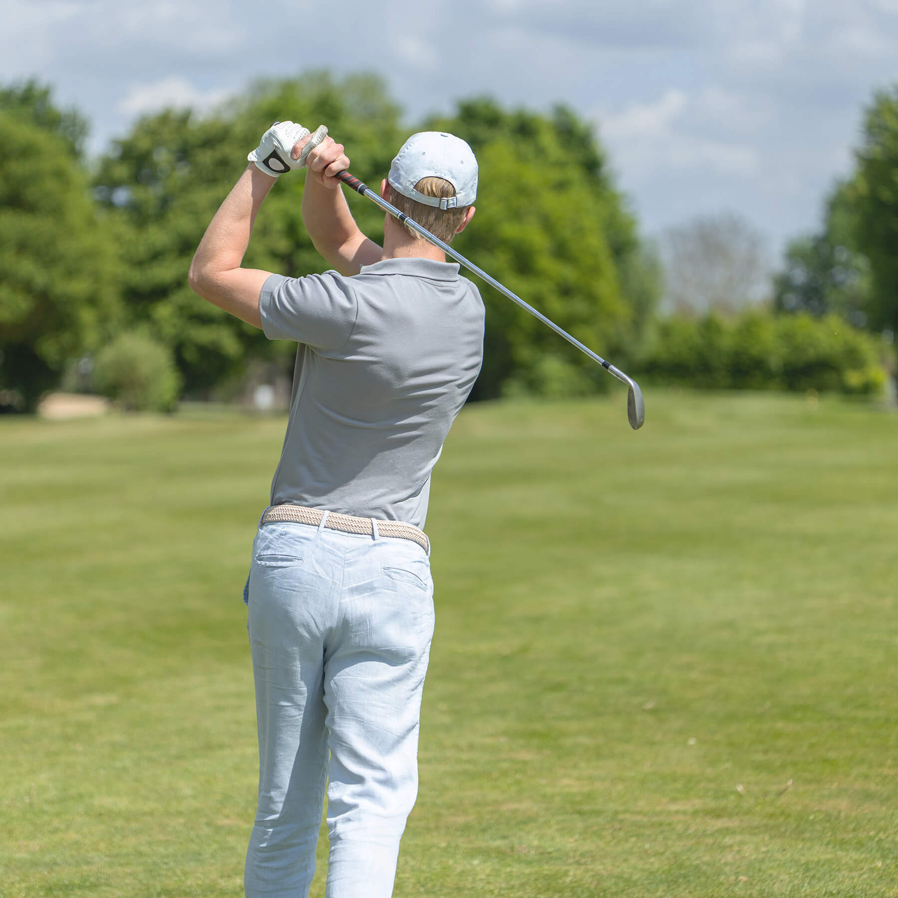 Golfspieler im grauen Lindenmann Poloshirt und beigem Stretchgürtel von Lindenmann führt einen Golfschwung auf dem Platz aus.