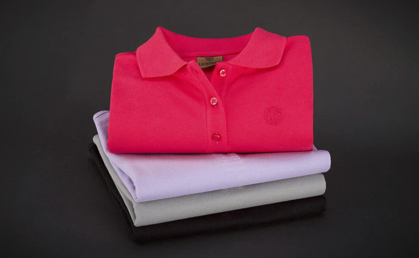 Drei gefaltete Poloshirts in Pink, Lila und Grau auf dunklem Hintergrund