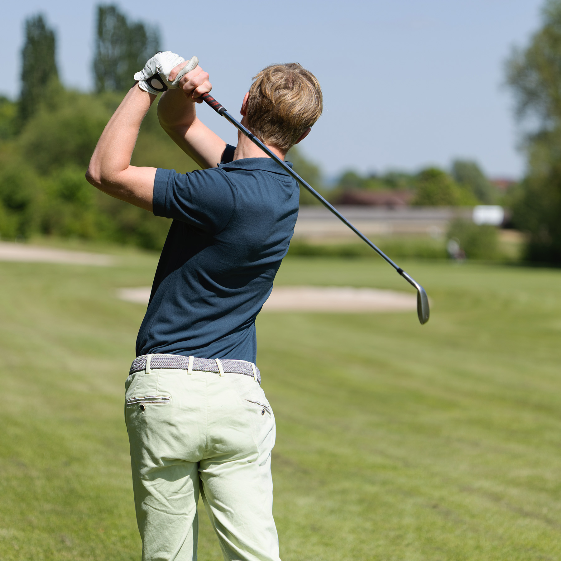 Feld, Natur, Im Freien, Person, Golf, Golfspieler im dunkelblauen Lindenmann Poloshirt und grauem Stretchgürtel führt einen Golfschwung auf dem Platz aus.